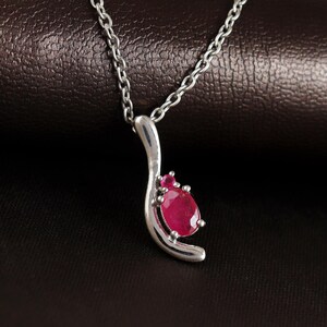 May include: A silver pendant necklace featuring a teardrop-shaped, vibrant red gemstone. The pendant has a curved, modern design with a smaller red stone accent. The chain is a delicate silver link.