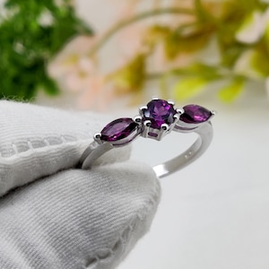 May include: A silver ring with three purple gemstones. The central stone is round, with two marquise-cut stones either side. The ring is set against a soft, blurred background, highlighting the jewellery's design and colour.