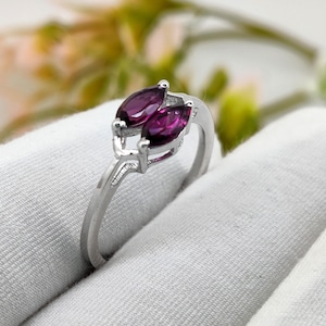May include: A silver ring with two marquise-cut, deep purple gemstones. The gemstones are set side-by-side in a prong setting, with a delicate silver band. The ring is displayed on a white surface, highlighting the ring's design and colour.