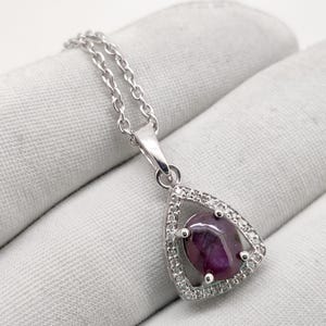 Op de afbeelding: Een zilveren hangerketting met een druppelvormige, dieppaarse edelsteen. De edelsteen is omgeven door kleine, heldere stenen en de hanger hangt aan een fijne zilveren ketting. De ketting wordt gepresenteerd op een wit oppervlak.