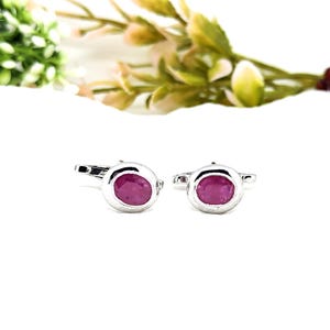 May include: A pair of silver cufflinks, each featuring an oval, deep pink gemstone. The cufflinks are set against a white background, with a blurred floral arrangement in the background.