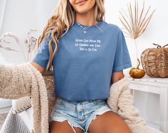 May include: A blue short-sleeved t-shirt with the text "When God Made Me, He Grinned and Said, 'This'll Be Fun'" in white. The shirt is paired with denim shorts. A beige sweater is draped over the shoulders.