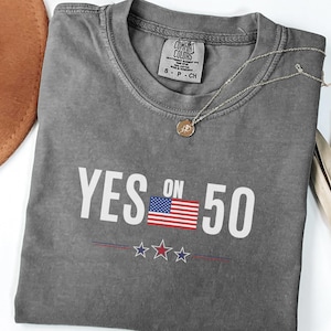 May include: A gray t-shirt with the text "YES ON 50" in white, featuring an American flag graphic. Below the text are three stars. A gold necklace and a brown hat are also visible.