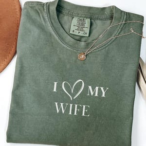 May include: Olive green t-shirt with the text "I ♡ MY WIFE" in white. A gold necklace with a pendant is on the shirt. A brown hat and dried plant are in the background.