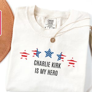 May include: Cream-colored t-shirt with the text "CHARLIE KIRK IS MY HERO" in black. Above the text are five stars, alternating between red and blue, with a design reminiscent of the American flag. A gold necklace rests on the shirt.