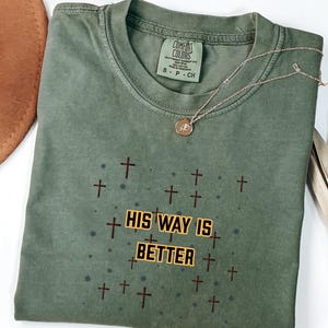 May include: Olive green t-shirt with the text "HIS WAY IS BETTER" in a yellow and black font, surrounded by brown crosses. A gold necklace with a pendant and a brown hat are also visible.