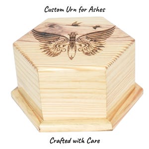 May include: A light-coloured wooden urn with a hexagonal shape. The top features an engraved design of wings and birds. The text "Custom Urn for Ashes" and "Crafted with Care" are visible on the image.