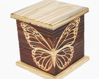 Puede incluir: Una caja de madera con un diseño de mariposa tallado en la parte delantera. La caja es de madera marrón oscuro con una mariposa marrón claro y una tapa marrón claro. La caja es rectangular con una tapa inclinada.