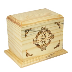 Könnte beinhalten: Eine hellfarbene Holzkiste mit einem eingravierten keltischen Kreuz auf der Vorderseite. Die rechteckige Kiste hat einen Sockel und dekorative Eckakzente. Die Holzmaserung ist sichtbar.