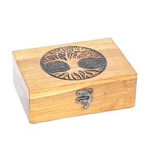 Puede incluir: Una caja de madera rectangular con un diseño de árbol de la vida tallado en la tapa. El árbol está dentro de un círculo oscuro. La caja tiene un cierre y bisagras de metal. La madera es de color marrón claro.