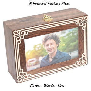May include: A rectangular wooden urn with a photo frame. The frame has an ornate white design. The urn is brown with a gold clasp. Text on the image reads "A Peaceful Resting Place" and "Custom Wooden Urn."