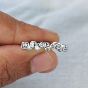 Op de afbeelding: Een zilveren ring met een uniek ontwerp met een reeks kleine, onregelmatig gevormde cirkels, elk versierd met een sprankelende, heldere edelsteen. De ring wordt tegen een zachte, witte achtergrond gehouden.