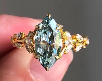 May include: A gold ring featuring a large, light blue marquise-cut gemstone as the centerpiece. The band is adorned with smaller, clear gemstones and leaf-like details, creating an elegant and nature-inspired design. The ring is held up against a neutral background.