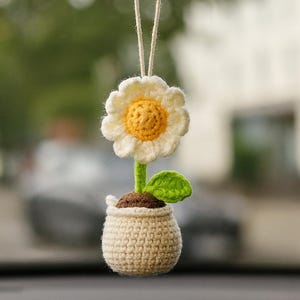 May include: A crocheted flower car charm. The charm features a white daisy with a yellow center, green stem and leaf, and a beige pot. The charm hangs from a string, perfect for adding a touch of handmade charm to any vehicle.