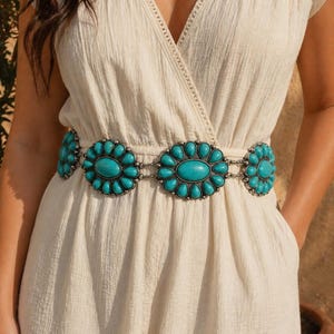 May include: A turquoise and silver-tone belt with a floral design is worn over a cream-colored jumpsuit. The belt features large turquoise stones set in a silver metal frame, connected by a chain.