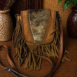 Puede incluir: Bolso bandolera de cuero marrón con cierre de solapa, adornado con flecos y un panel central de piel de animal. El bolso presenta detalles de tachuelas y una correa larga y ajustable. El bolso mide aproximadamente 25 cm de ancho.