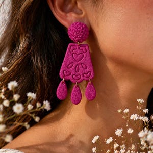 Puede incluir: Primer plano de un pendiente magenta. El pendiente tiene una bola texturizada en la parte superior, una sección media rectangular con recortes de corazón y flores, y tres colgantes en forma de lágrima. Flores blancas en el fondo.