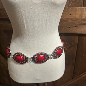 May include: A silver-toned metal belt with large, oval-shaped red stones. The stones are set in a decorative frame and connected by a chain. The belt is displayed on a mannequin.