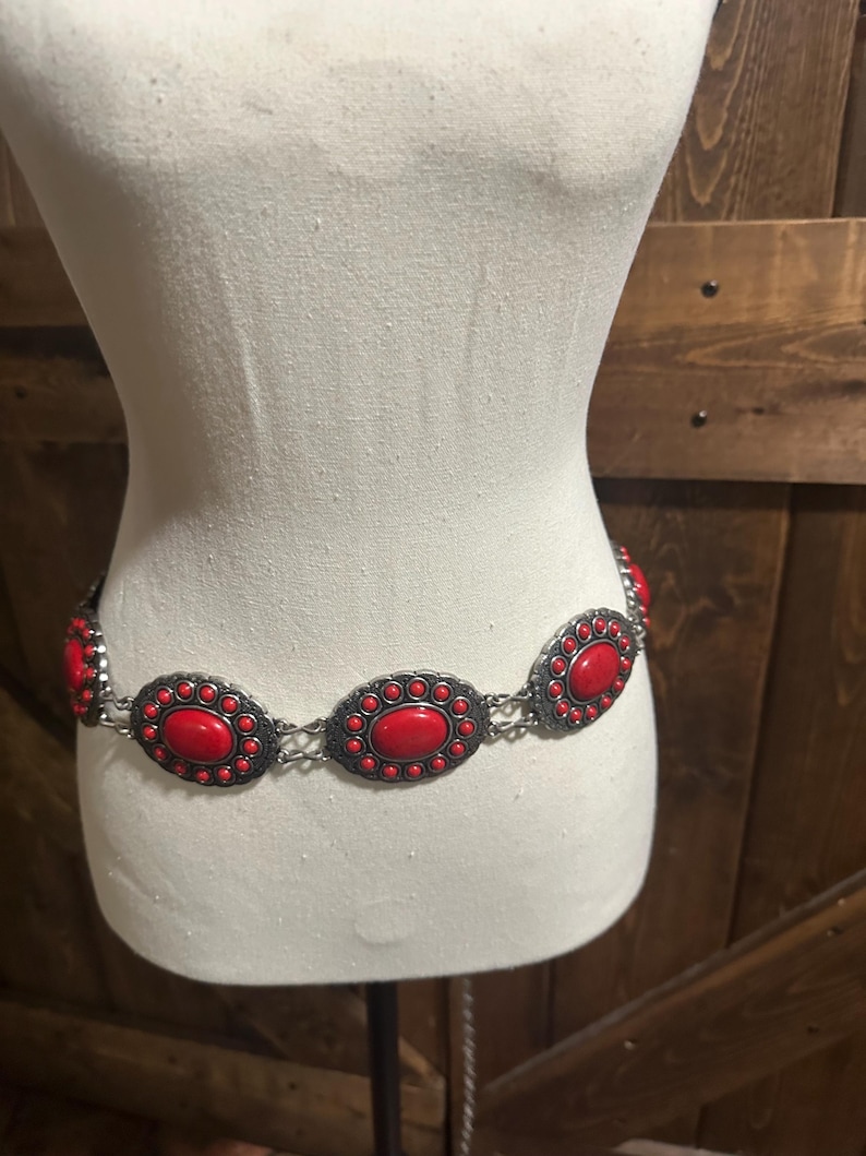 May include: A silver-toned metal belt with large, oval-shaped red stones. The stones are set in a decorative frame and connected by a chain. The belt is displayed on a mannequin.