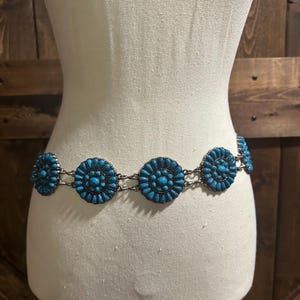 May include: A turquoise and silver-tone belt with circular turquoise embellishments. The belt is displayed on a mannequin, showcasing its design and style. The turquoise stones are arranged in a floral pattern.