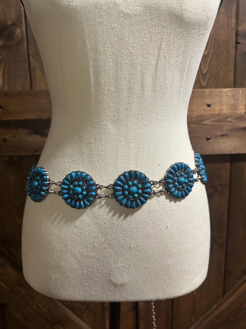 May include: A turquoise and silver-tone belt with circular turquoise embellishments. The belt is displayed on a mannequin, showcasing its design and style. The turquoise stones are arranged in a floral pattern.