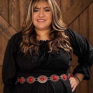 May include: A woman wearing a black dress with a decorative belt. The belt is silver-toned with large, oval, red stones. The woman has long, wavy hair with blonde highlights. The background is a wooden door.