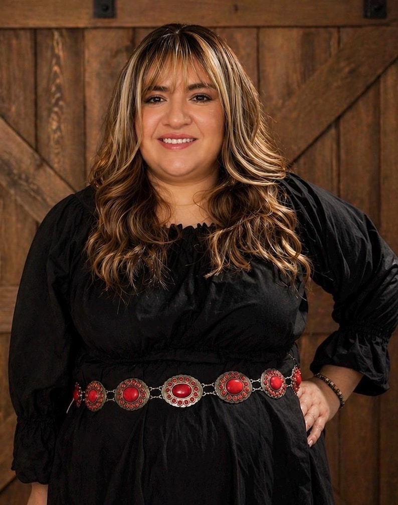 May include: A woman wearing a black dress with a decorative belt. The belt is silver-toned with large, oval, red stones. The woman has long, wavy hair with blonde highlights. The background is a wooden door.