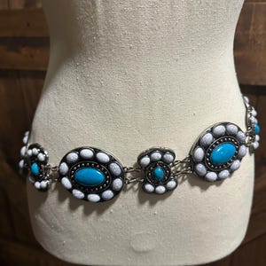 May include: A silver-toned belt with turquoise and white oval and floral-shaped embellishments. The belt is displayed on a mannequin and features a series of connected decorative elements. The turquoise stones are the focal point, surrounded by white and silver accents.