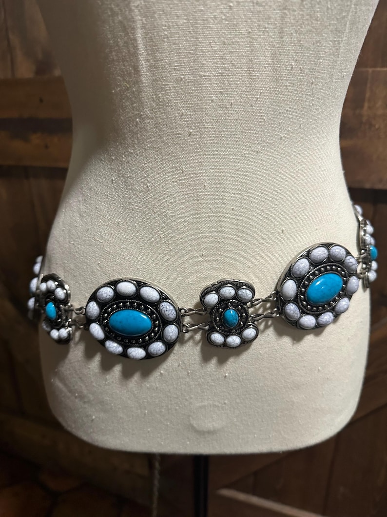 May include: A silver-toned belt with turquoise and white oval and floral-shaped embellishments. The belt is displayed on a mannequin and features a series of connected decorative elements. The turquoise stones are the focal point, surrounded by white and silver accents.