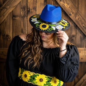 Puede incluir: Un sombrero fedora azul con ala negra, adornado con girasoles amarillos bordados y calas blancas. El sombrero se usa con un vestido negro con hombros descubiertos y un cinturón bordado con girasoles amarillos. El fondo es una puerta de madera.
