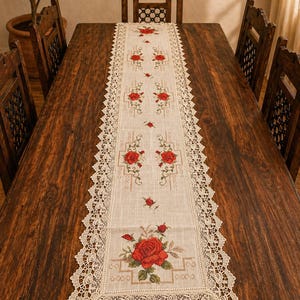 Puede incluir: Un camino de mesa rectangular de color crema con un diseño floral. El camino de mesa presenta rosas rojas bordadas y hojas verdes, enmarcadas por un delicado borde de encaje festoneado. El camino de mesa está colocado sobre una mesa de madera oscura.