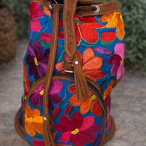 May include: A blue embroidered backpack with a floral design in shades of red, orange, purple, and pink. The backpack has brown leather straps and trim, a drawstring closure, and a zippered front pocket.