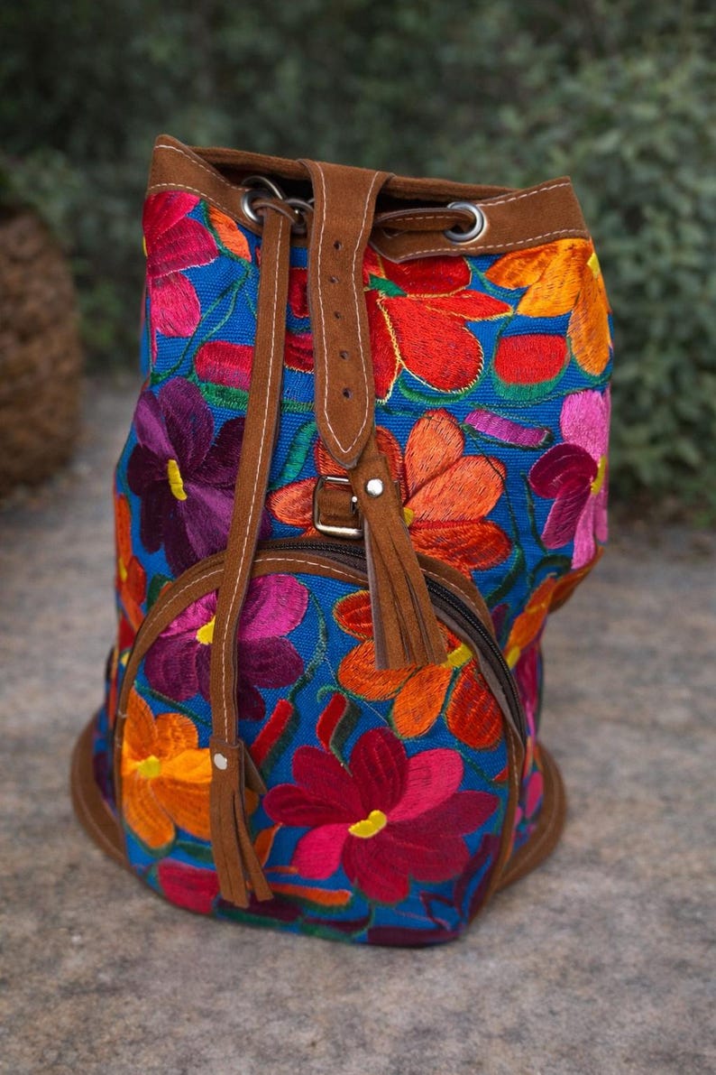 May include: A blue embroidered backpack with a floral design in shades of red, orange, purple, and pink. The backpack has brown leather straps and trim, a drawstring closure, and a zippered front pocket.