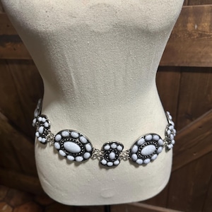 May include: A silver-toned belt with white oval and circular embellishments. The belt is displayed on a mannequin, showcasing its design. The white stones are set in a black border, creating a striking contrast. The belt is a fashion accessory.
