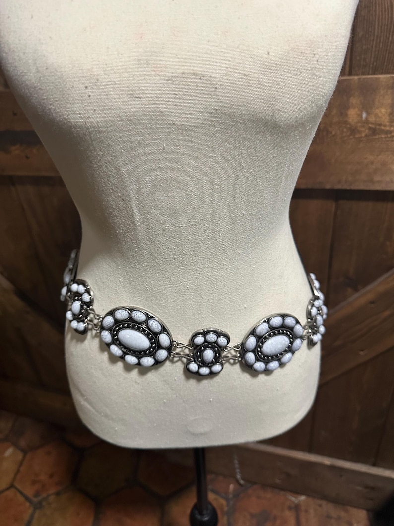 May include: A silver-toned belt with white oval and circular embellishments. The belt is displayed on a mannequin, showcasing its design. The white stones are set in a black border, creating a striking contrast. The belt is a fashion accessory.