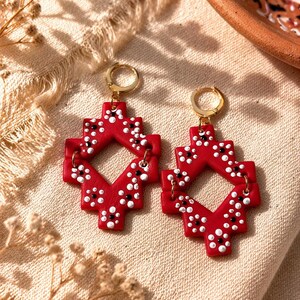May include: A pair of red geometric earrings with gold-colored hoops. The earrings feature a cross-like design with white and black dot details. The earrings are displayed on a beige fabric.