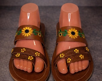 May include: Brown leather sandals with embroidered yellow sunflowers and green leaves on the straps. The sandals have a toe loop and a strap across the top of the foot, featuring small yellow flower details. The soles are brown.