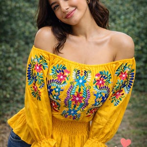 May include: A yellow off-the-shoulder top with long sleeves, adorned with colourful floral embroidery in pink, blue, and purple. The top has a ruffled hem and the word "Coqueta" in cursive.