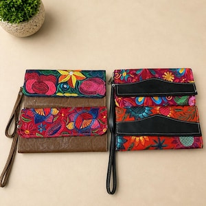May include: Two embroidered clutch wallets with wrist straps. One is brown with red, pink, and yellow floral embroidery. The other is black with red, orange, and blue floral embroidery. Both have black wrist straps.