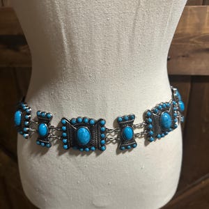 May include: A silver-toned belt with turquoise-colored stones. The belt features rectangular and oval-shaped decorative elements connected by small chains. The turquoise stones are set within a detailed, ornate design.