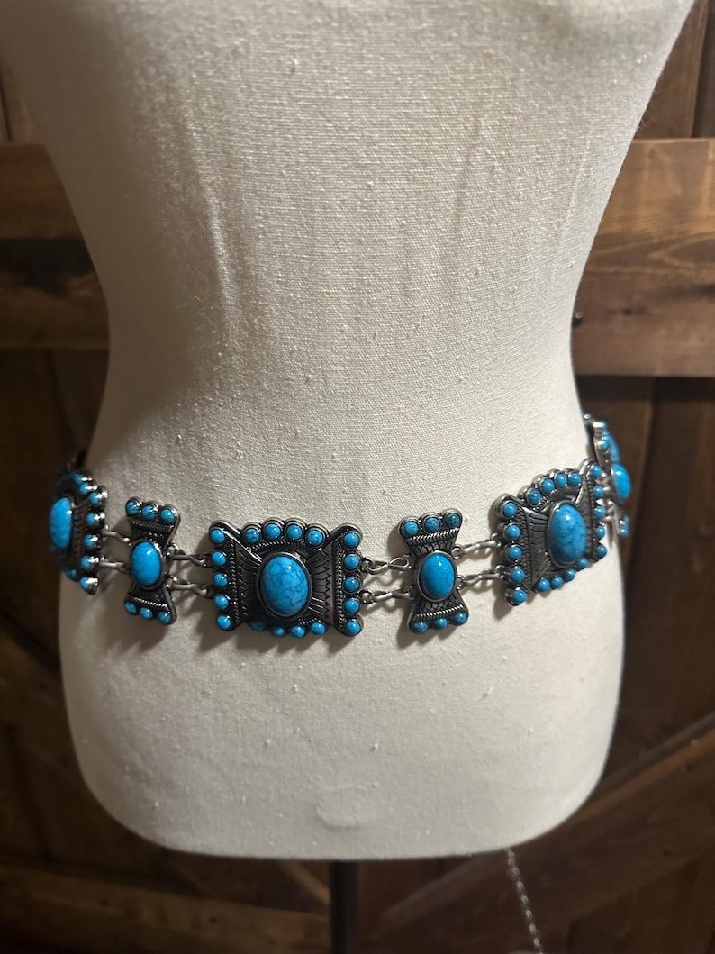 May include: A silver-toned belt with turquoise-colored stones. The belt features rectangular and oval-shaped decorative elements connected by small chains. The turquoise stones are set within a detailed, ornate design.