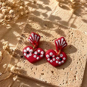 Puede incluir: Un par de pendientes rojos hechos a mano con forma de corazón y detalles de puntos blancos y negros. Los pendientes tienen un aro dorado y un acento blanco en forma de concha. Se muestran sobre una superficie beige texturizada.