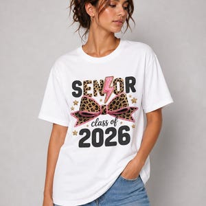 Puede incluir: Camiseta blanca con el texto "SENIOR class of 2026". El diseño incluye un rayo rosa, un lazo con estampado de leopardo y estrellas doradas. La camiseta es de cuello redondo.