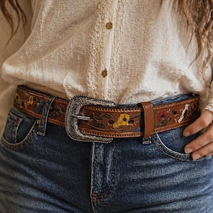 May include: A brown leather belt with floral and bird designs, worn with blue jeans. The belt features a large, ornate silver buckle. The belt is decorated with yellow, blue, and red flowers.