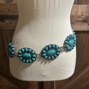 May include: A silver-toned belt with turquoise-colored oval and floral-shaped embellishments. The belt is displayed on a mannequin, showcasing its design and style. The turquoise stones are set in a silver metal frame.