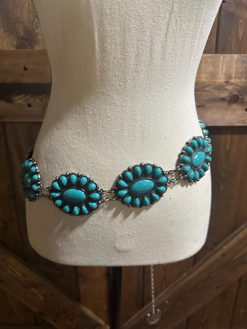May include: A silver-toned belt with turquoise-colored oval and floral-shaped embellishments. The belt is displayed on a mannequin, showcasing its design and style. The turquoise stones are set in a silver metal frame.