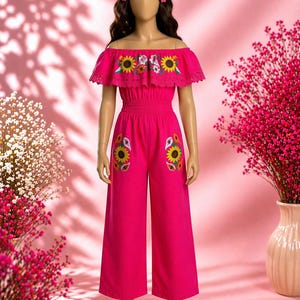 Puede incluir: Un mono rosa vibrante con bordado de girasoles. La parte superior con hombros descubiertos tiene un borde con volantes y los pantalones tienen diseños de girasoles en los bolsillos. El atuendo se exhibe en un maniquí con pinzas para el pelo de flores.