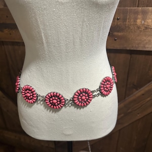 May include: A pink floral belt with a silver chain. The belt is displayed on a mannequin. The pink flowers are evenly spaced around the belt. The background is a wooden door.