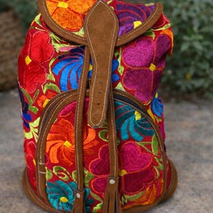 May include: A vibrant, embroidered backpack with a red background and colorful floral patterns in orange, pink, blue, and green. It features brown leather accents, a flap closure, and adjustable straps. The bag has a round base and a drawstring closure.
