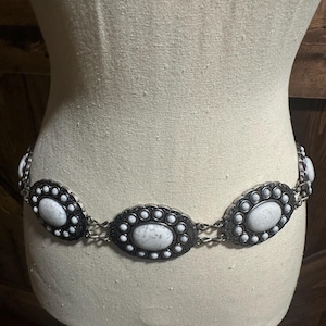 May include: A silver-toned belt with oval, white stone-like embellishments. Each oval is framed by a black border and surrounded by smaller white accents. The belt is displayed on a mannequin.